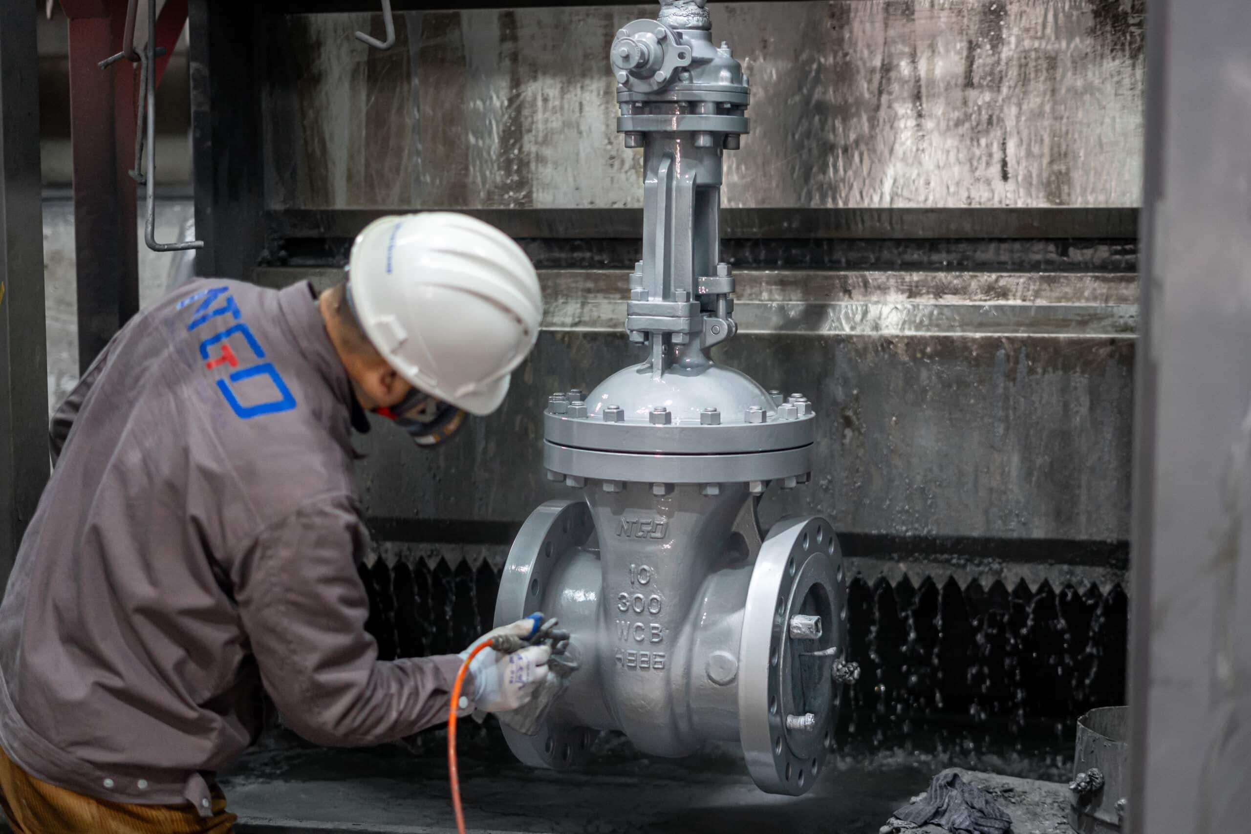 Un trabajador inspecciona o aplica un recubrimiento a una válvula de compuerta industrial en el taller de NTGD.